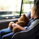 Ein Mädchen sitzt in einem Kindersitz im Auto und schläft. Sie hält einen Teddybären dabei.