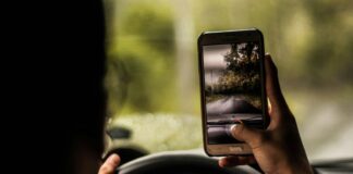 Eine Person fährt auf einer Landstraße im Auto. Sie hält ein Smartphone in der rechten Hand, der Daumen am Auslöser der Kamera. Die linke Hand ruht am Steuer.