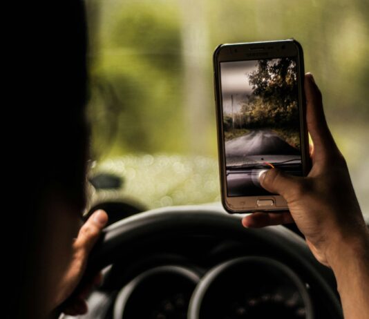 Eine Person fährt auf einer Landstraße im Auto. Sie hält ein Smartphone in der rechten Hand, der Daumen am Auslöser der Kamera. Die linke Hand ruht am Steuer.