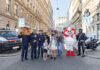 Auf einem Zebrastreifen mit Schulweg-Tafeln stehen Polizeibär, HELMI, Kinder und Polizisten sowie KFV-Expert*innen. Sie strahlen in die Kamera.