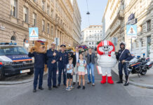 Auf einem Zebrastreifen mit Schulweg-Tafeln stehen Polizeibär, HELMI, Kinder und Polizisten sowie KFV-Expert*innen. Sie strahlen in die Kamera.