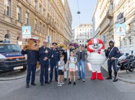 Auf einem Zebrastreifen mit Schulweg-Tafeln stehen Polizeibär, HELMI, Kinder und Polizisten sowie KFV-Expert*innen. Sie strahlen in die Kamera.