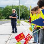 Eine Polizeibeamtin, die mit Kindern an einer Straße stehen. Die Kinder sich ausgestattet mit gelben Warnwesten, Fahnen und Klappbrettern und stehen aufgereiht am Straßenrand.