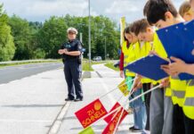 Eine Polizeibeamtin, die mit Kindern an einer Straße stehen. Die Kinder sich ausgestattet mit gelben Warnwesten, Fahnen und Klappbrettern und stehen aufgereiht am Straßenrand.