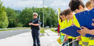 Eine Polizeibeamtin, die mit Kindern an einer Straße stehen. Die Kinder sich ausgestattet mit gelben Warnwesten, Fahnen und Klappbrettern und stehen aufgereiht am Straßenrand.