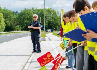 Eine Polizeibeamtin, die mit Kindern an einer Straße stehen. Die Kinder sich ausgestattet mit gelben Warnwesten, Fahnen und Klappbrettern und stehen aufgereiht am Straßenrand.