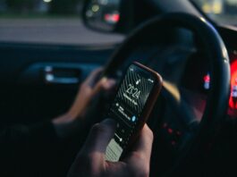 Man sieht Hände einer Person, nachts im Auto, mit einer Hand hält sie das Lenkrad, mit der anderen tipps sie am Handy herum.