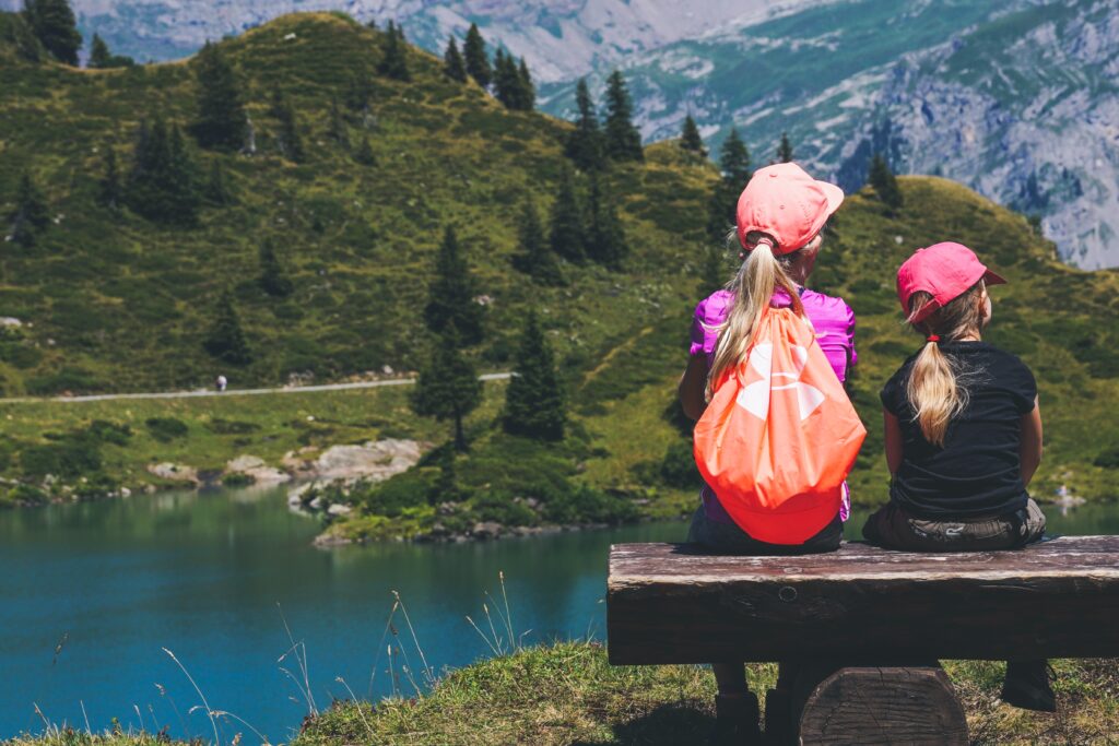 Zwei Kinder sitzen auf einer Bank und schauen in eine Berglandschaft mit See, Wald und Gebirgsrücken. Die beiden Mädchen werden von hinten gezeigt und tragen beide eine Kappe, die größere hat auch einen Rucksack/Beutel umgeschnallt.